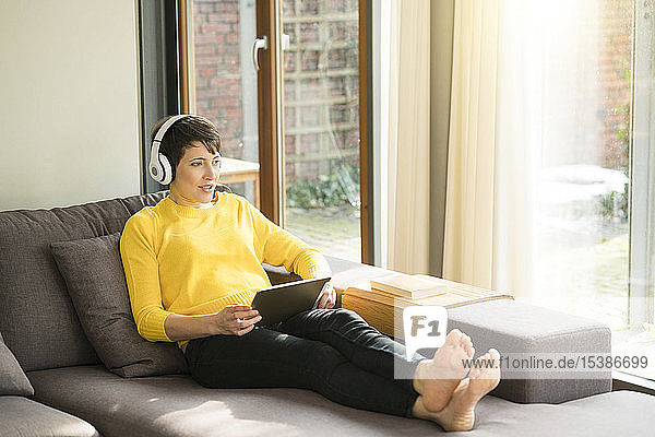 Portrait of woman with digital tablet sitting on the couch at home listening music with headphones