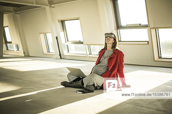 Pregnant busnesswoman sitting on floor of new office rooms  using VR goggles and laptop