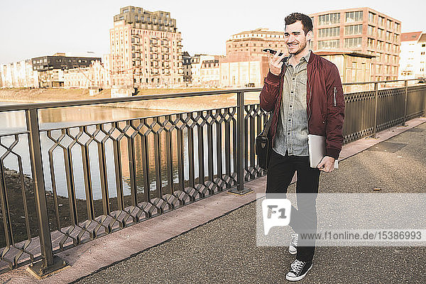 Smiling young man with tablet  cell phone and bag on a bridge in the city