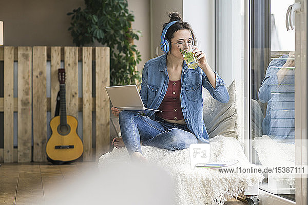 Woman with headphones and laptop sitting at the window at home drinking from glass