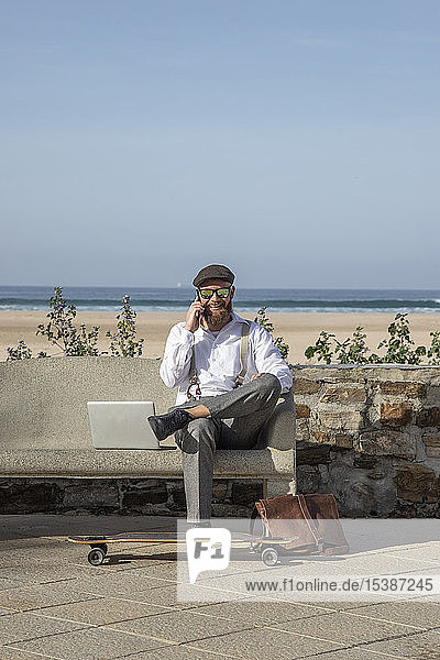 Lächelnder Mann am Telefon  der mit Laptop auf einer Bank vor Strand und Meer sitzt