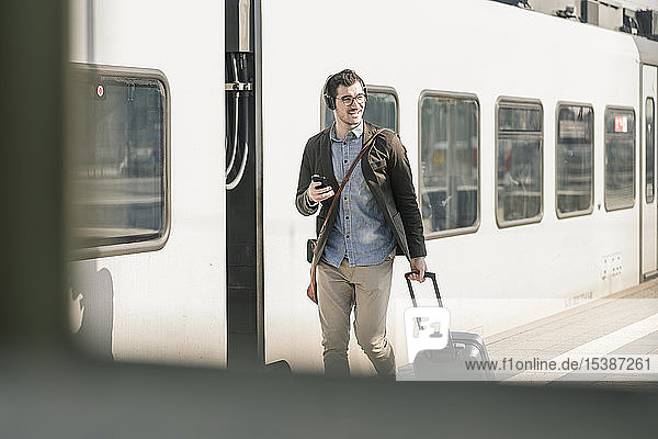 Lächelnder junger Mann mit Kopfhörern Handy und Koffer auf dem Bahnsteig