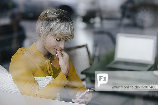 Blonde Geschäftsfrau sitzt am Fenster und bezahlt mit Smartphone und Kreditkarte