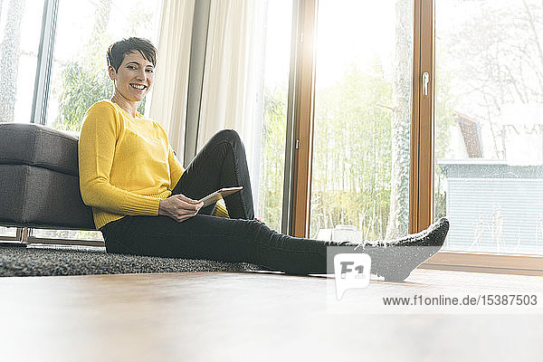 Porträt einer glücklichen Frau mit digitalem Tablett  die auf dem Boden des Wohnzimmers sitzt
