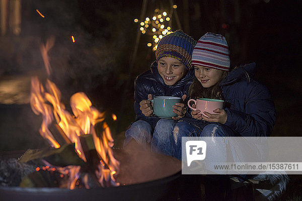 Junge und Mädchen mit Bechern sitzen nachts am Feuer