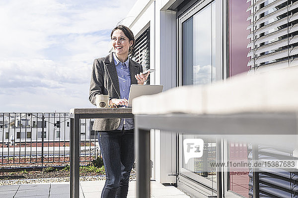 Lächelnde Geschäftsfrau mit Handy und Laptop auf der Dachterrasse