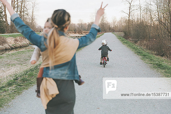 Mutter trägt Tochter und jubelt ihrem kleinen Sohn zu  fährt Fahrrad