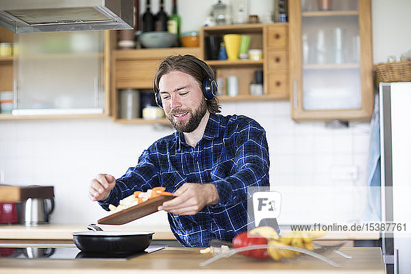 Junger Mann mit Bart und kariertem Hemd und Kopfhörer  der in der Küche Gemüse kocht