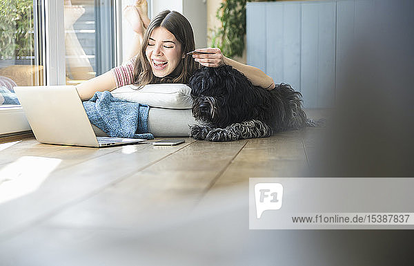 Glückliche junge Frau mit Hund am Fenster liegend beim Online-Shopping zu Hause