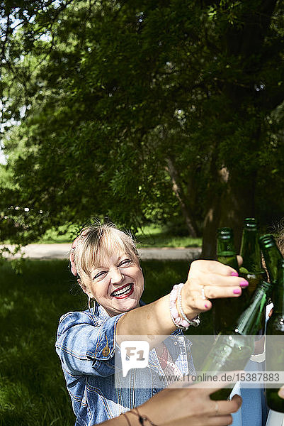 Porträt einer glücklichen Frau  die mit Freunden im Park an einer Bierflasche klimpert