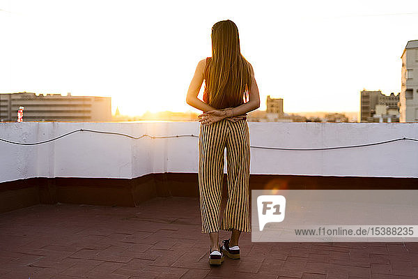 Rückansicht eines jugendlichen Mädchens  das bei Sonnenuntergang auf einer Dachterrasse in der Stadt steht