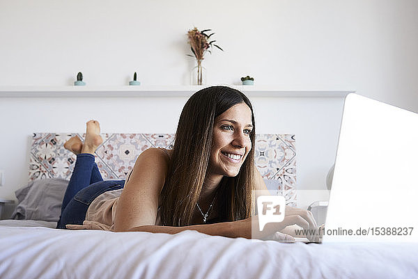Porträt einer lächelnden jungen Frau  die mit einem Laptop auf dem Bett liegt