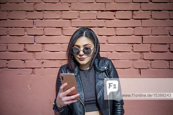 Porträt einer jungen Frau mit Sonnenbrille und schwarzer Lederjacke  die ein Smartphone betrachtet