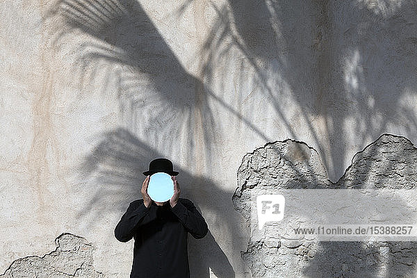 Morocco  Essaouira  man wearing a bowler hat holding mirror in front of his face at a wall