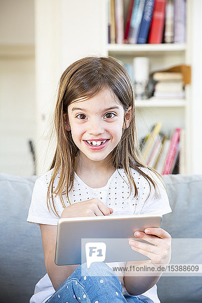 Portrait of happy little girl sitting on the couch at home using digital tablet