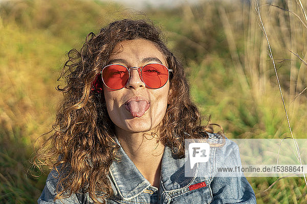 Porträt einer jungen Frau mit lockigem braunem Haar und roter Sonnenbrille  die die Zunge herausstreckt