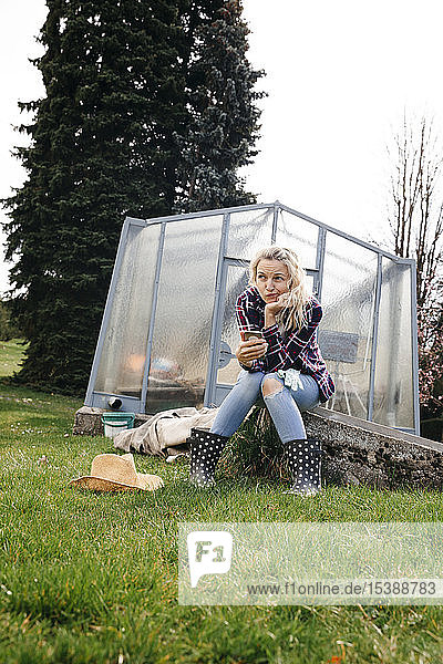 Junge Frau mit Strohhut sitzt vor einem Gewächshaus und hält ihr Mobiltelefon