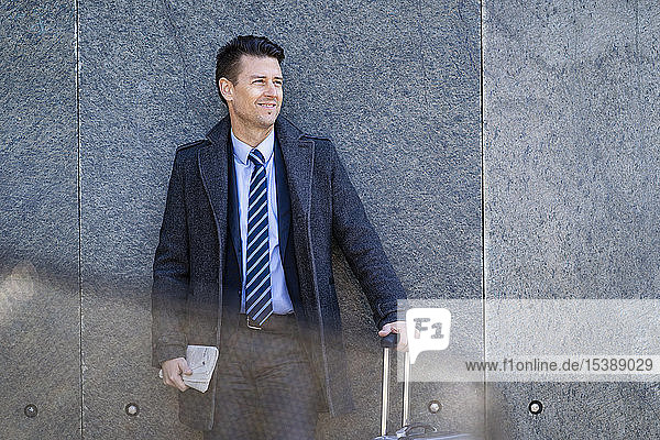 Smiling businessman with suitcase standing at a wall