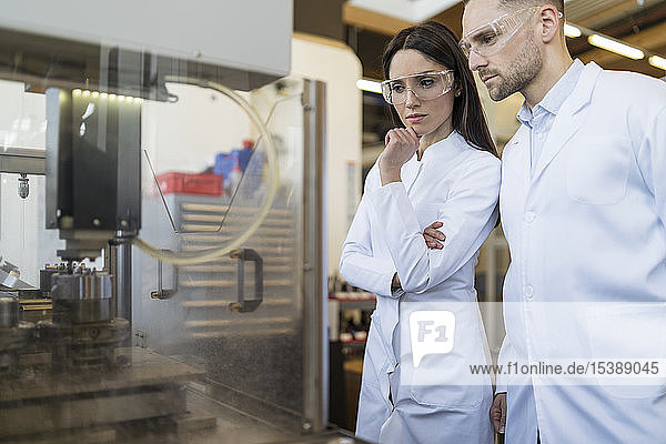 Colleagues wearing lab coats and safety goggles looking at machine in modern factory
