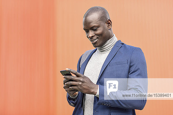 Lächelnder Geschäftsmann steht vor oranger Wand und schaut auf Smartphone