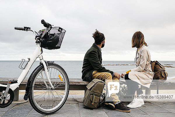 Couple sitting on a bench at beach promenade next to e-bike looking at the sea
