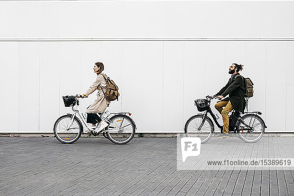 Couple riding e-bikes along a white wall in the city