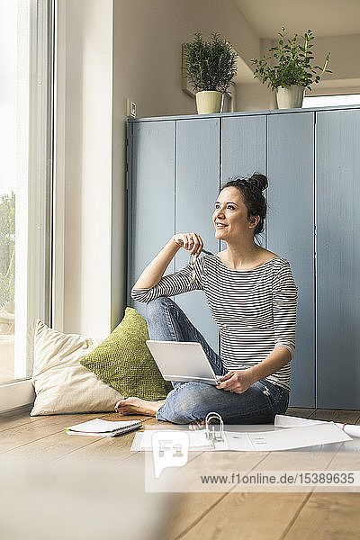 Lächelnde Frau sitzt zu Hause am Fenster und arbeitet mit Laptop und Dateiordner