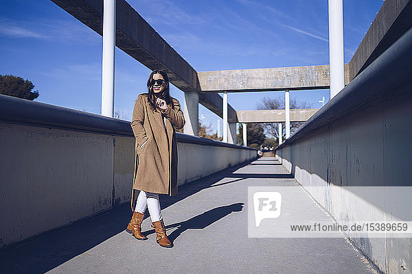 Smiling young woman wearing coat and sunglasses on a bridge