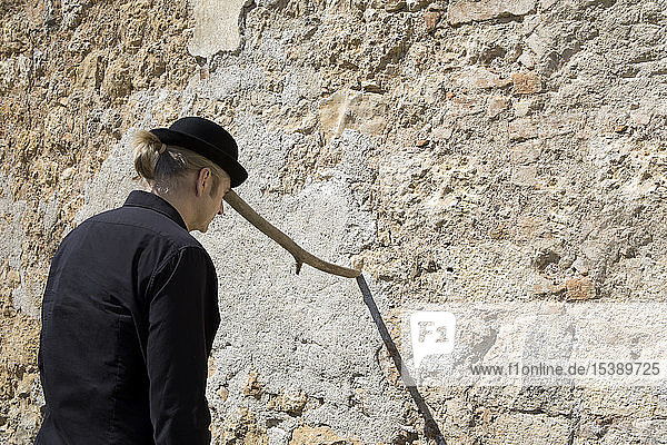 Man wearing a bowler hat balancing a stick at a stone wall