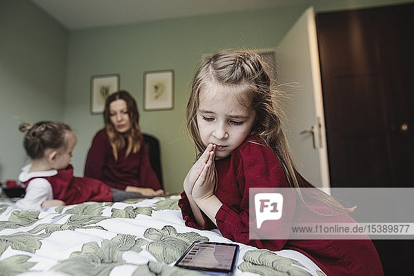 Mädchen schaut auf Smartphone im Bett mit Mutter und Schwester im Hintergrund