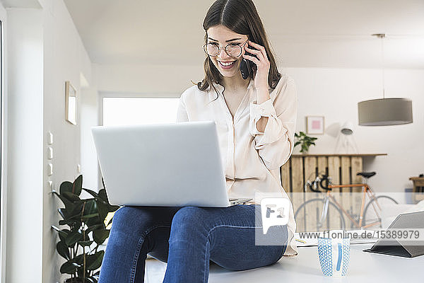 Smiling young woman sitting on table at home using laptop and cell phone