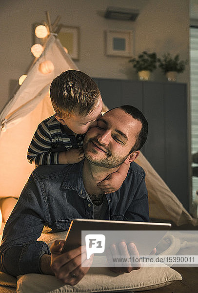 Son kissing father with tablet at an illuminated tent at home