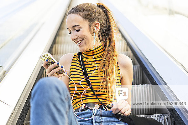 Teenager-Mädchen sitzt mit Kopfhörern und Smartphone auf der Rolltreppe