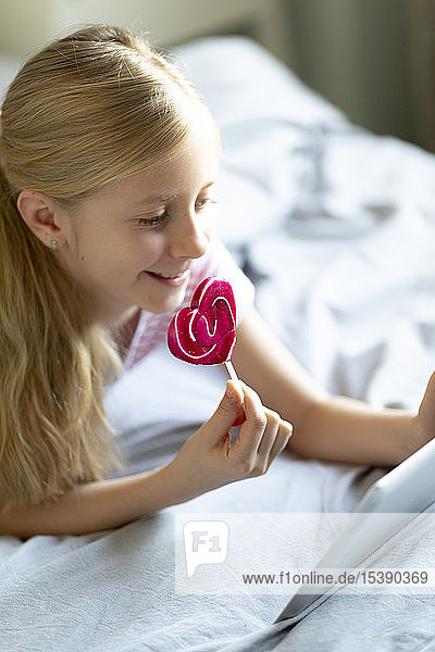 Lächelndes blondes Mädchen liegt mit einem Lolli auf dem Bett und benutzt ein digitales Tablett
