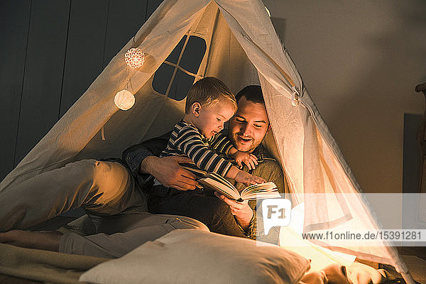 Father reading book to son at an illuminated tent at home