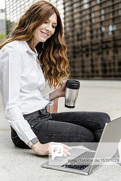 Young businesswoman sitting on bench in the city  working with laptop  drinking coffee