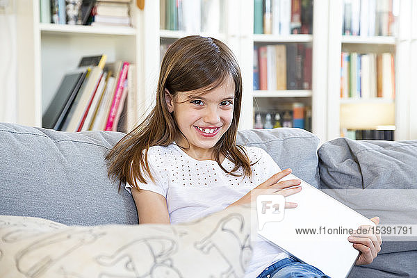 Porträt eines fröhlichen kleinen Mädchens  das zu Hause auf der Couch mit einem digitalen Tablet sitzt