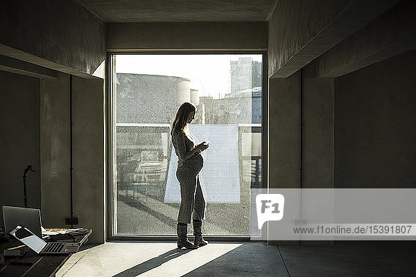 Pregnant businesswoman standing at office window  using smartphone