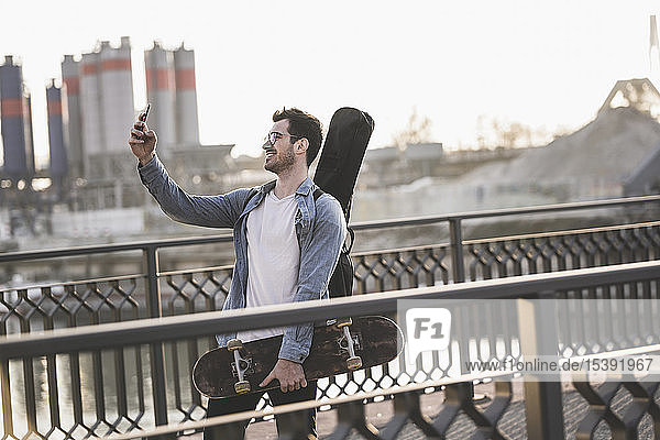 Junger Mann mit Gitarrenkoffer und Skateboard beim Selfie in der Stadt