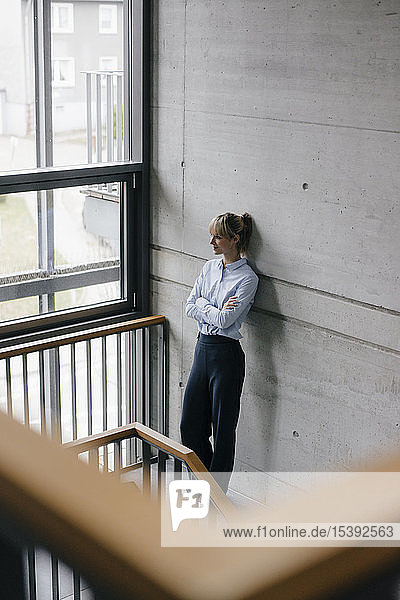 Successful businesswoman leaning on wall  looking out of window  with arms crossed
