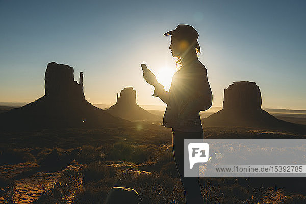 USA  Utah  Monument Valley  Silhouette einer Frau mit Cowboyhut  die bei Sonnenaufgang auf ein Mobiltelefon schaut