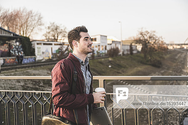 Young man with tablet  bag and takeaway coffee on a bridge in the city