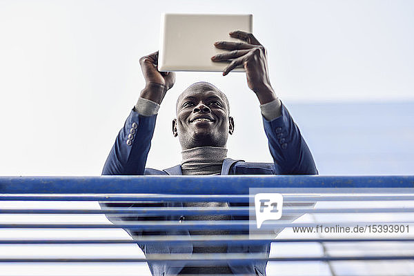 Porträt eines lächelnden Geschäftsmannes  der sich mit einem digitalen Tablett an ein Geländer lehnt