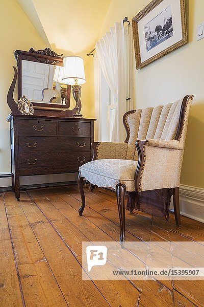 Antique sculpted wood and upholstered armchair dresser with mirror in guest bedroom with oil stained fir floorboards inside an old 1900 Victorian Queen Anne revival style house Quebec Canada. This image is property released.