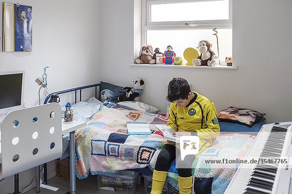 England Uk-Young boy 11 years in Little league football outfit reads a book in his bedroom.