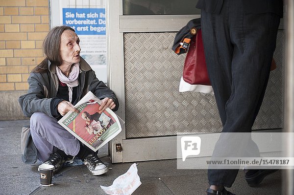 Berlin Germany. Homeless and impoverished mature adult woman sitting down on the pavement at Ostbahnhof trying to sell a newspaper and magazine helping her to earn a small living.
