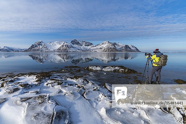 Man standing on a rock taking pictures of the Limstrandtinden snowcapped group in the background in winter. Vestvagoy municipality Lofoten district Nordland county Norway.