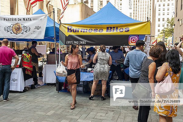 Tourists and office workers flock to Burmese Bites in The Outpost of the Queens Night Market located in Rockefeller Plaza in New York on Tuesday July 30 2019. The marketplace features ethnic and artisanal foods by small entrepreneurs all of whom are regulars at the Queens Night Market. Nine vendors will encamp in the plaza Monday through Thursday for three weeks. (© Richard B. Levine).