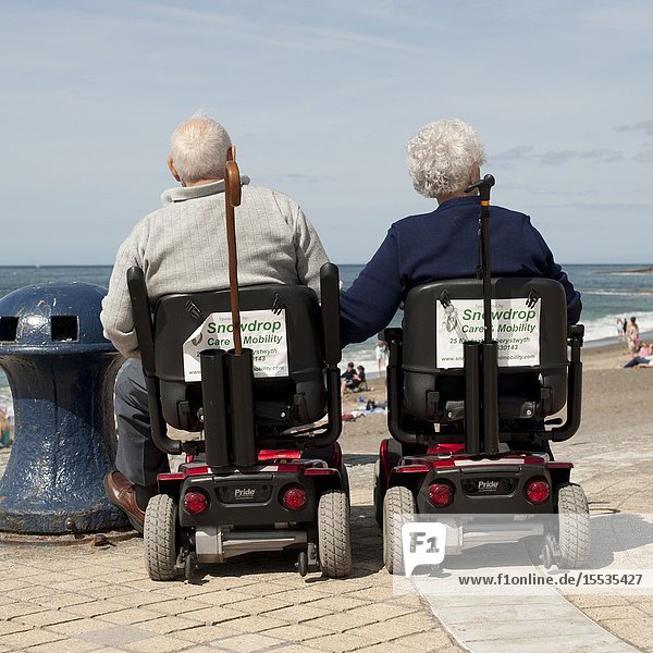 Rear view of an elderly couple husband and wife at the seaside sitting in powered mobility scooters Aberystwyth Wales UK.