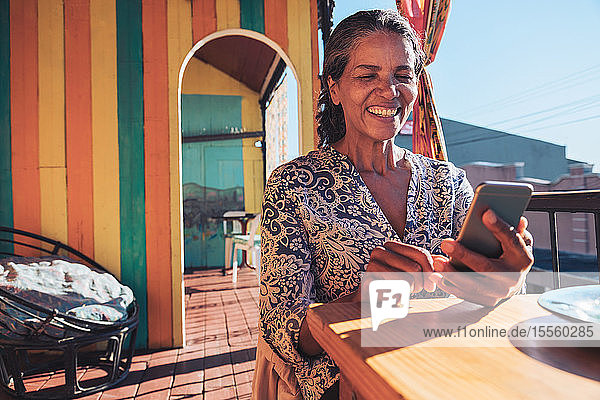 Lächelnde  glückliche Frau mit Smartphone auf dem Balkon eines sonnigen Restaurants